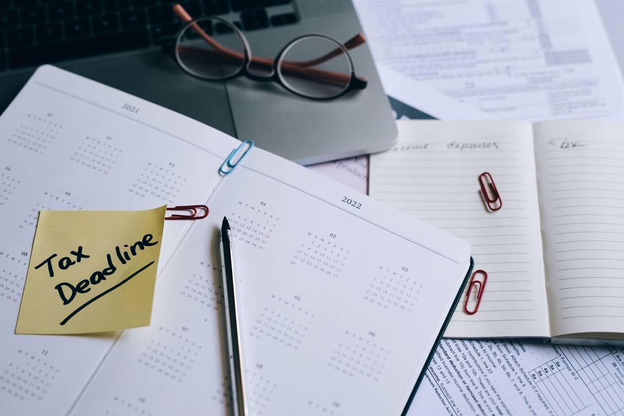 Cuốn sổ lịch mở trên bàn với tờ giấy nhớ màu vàng ghi Tax Deadline, bút, kẹp giấy và kính