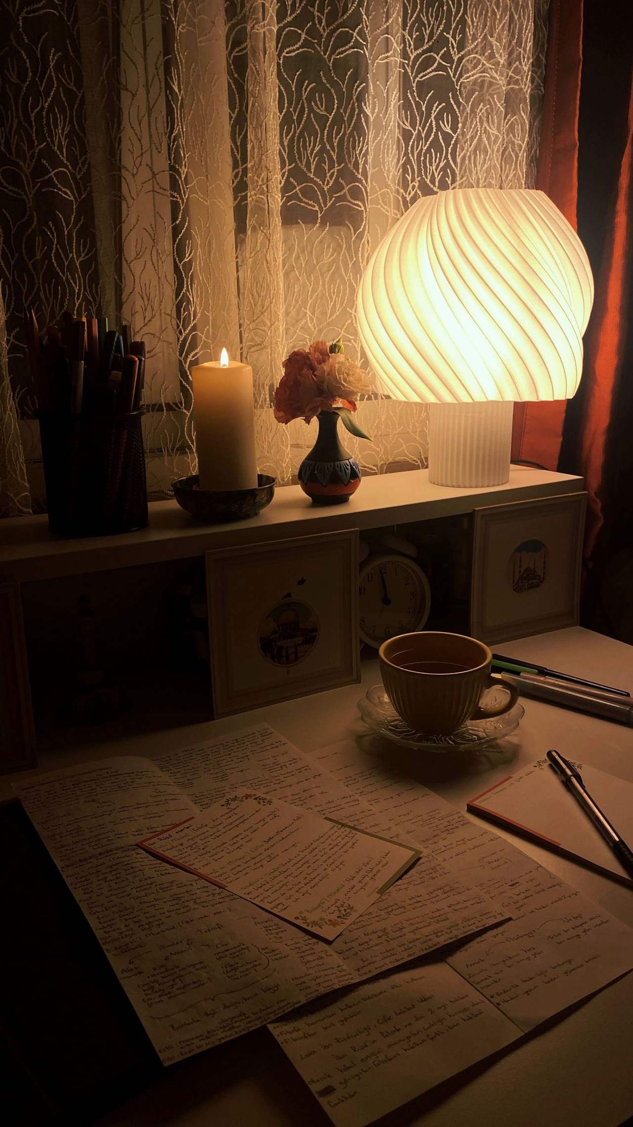 A desk in the evening lit by a lamp and a candle, with a cup of tea and handwritten notes