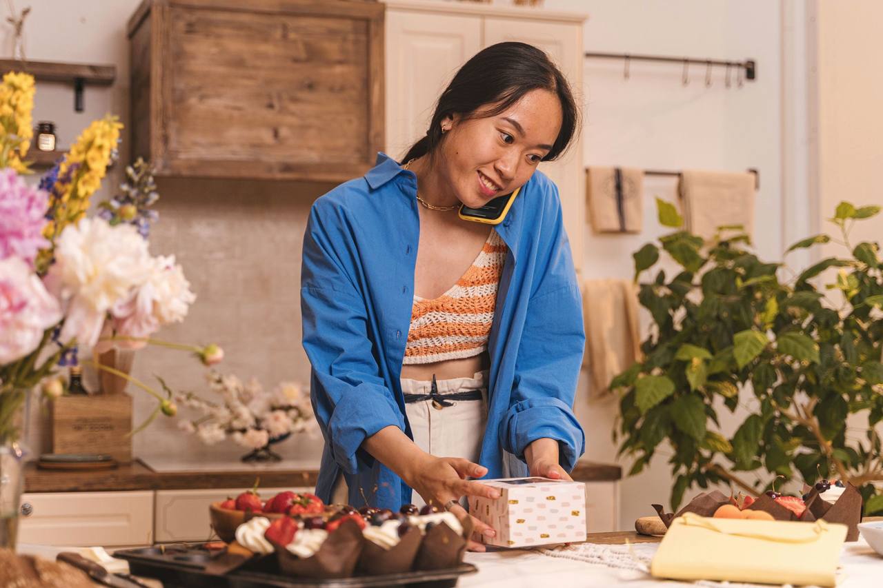 Người phụ nữ châu Á mặc áo sơ mi xanh vừa kẹp điện thoại nghe máy vừa đóng hộp bánh trên quầy, trước mặt là khay bánh kem phủ dâu tây và cherry