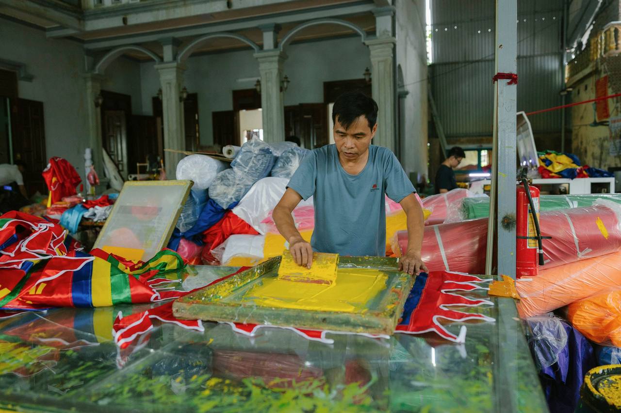 Người đàn ông trung niên mặc áo thun xanh xám đứng bên bàn in, tay kéo dao gạt mực vàng qua khung lưới, xung quanh là những cuộn hàng bọc nilon và tấm in nhiều màu chất trong xưởng