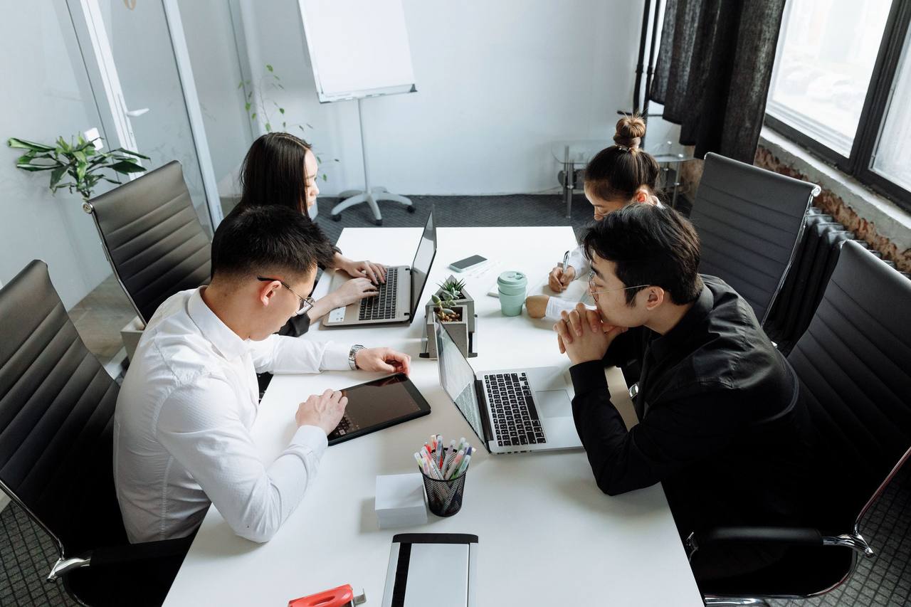 Bốn đồng nghiệp người châu Á ngồi quanh chiếc bàn trắng làm việc với máy tính xách tay và máy tính bảng trong một phòng làm việc nhỏ có bảng lật và cửa sổ lớn