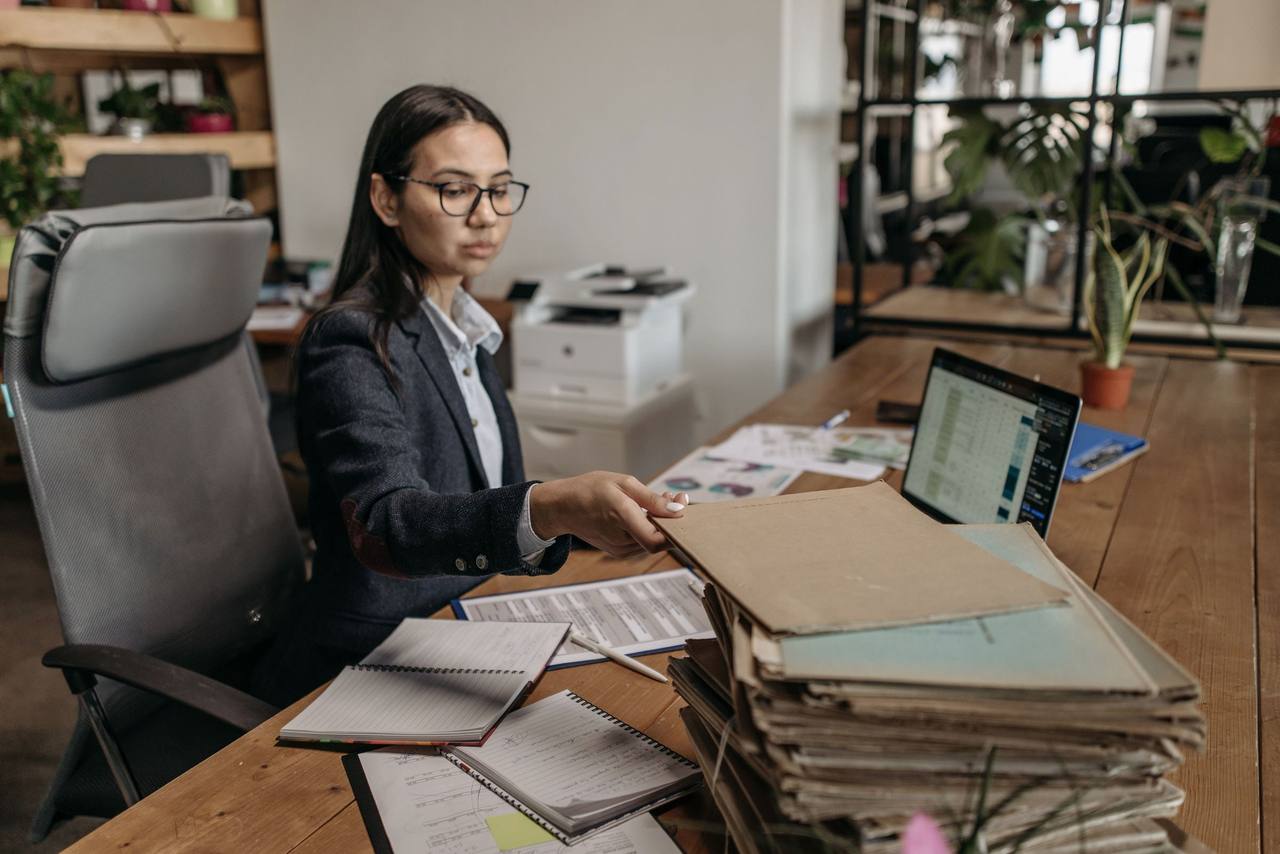 Nữ nhân viên văn phòng đeo kính làm việc bên máy tính xách tay và chồng bìa hồ sơ giấy trên bàn gỗ