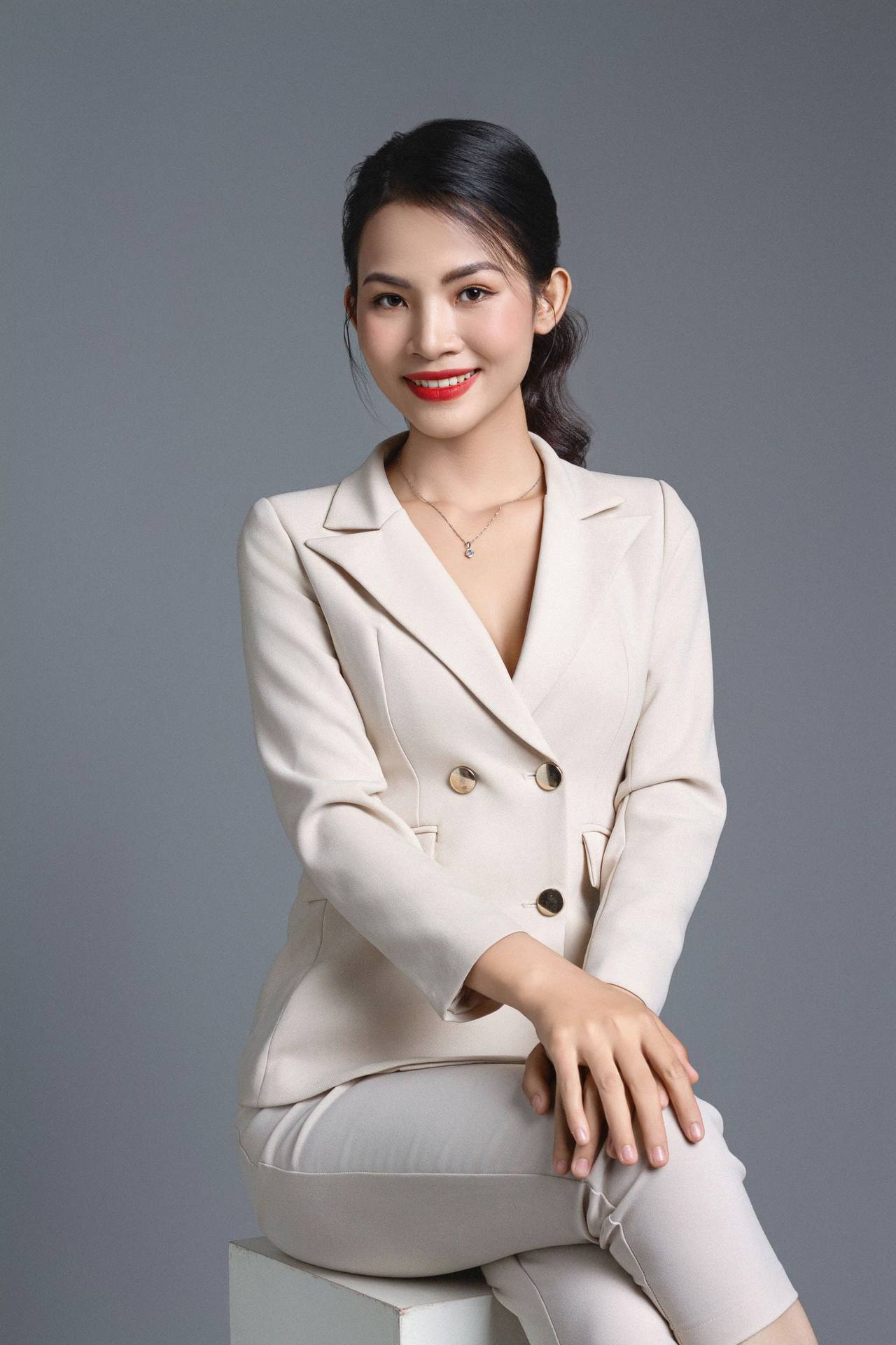 Chân dung người phụ nữ Việt Nam mặc bộ vest màu kem ngồi mỉm cười trước phông nền xám