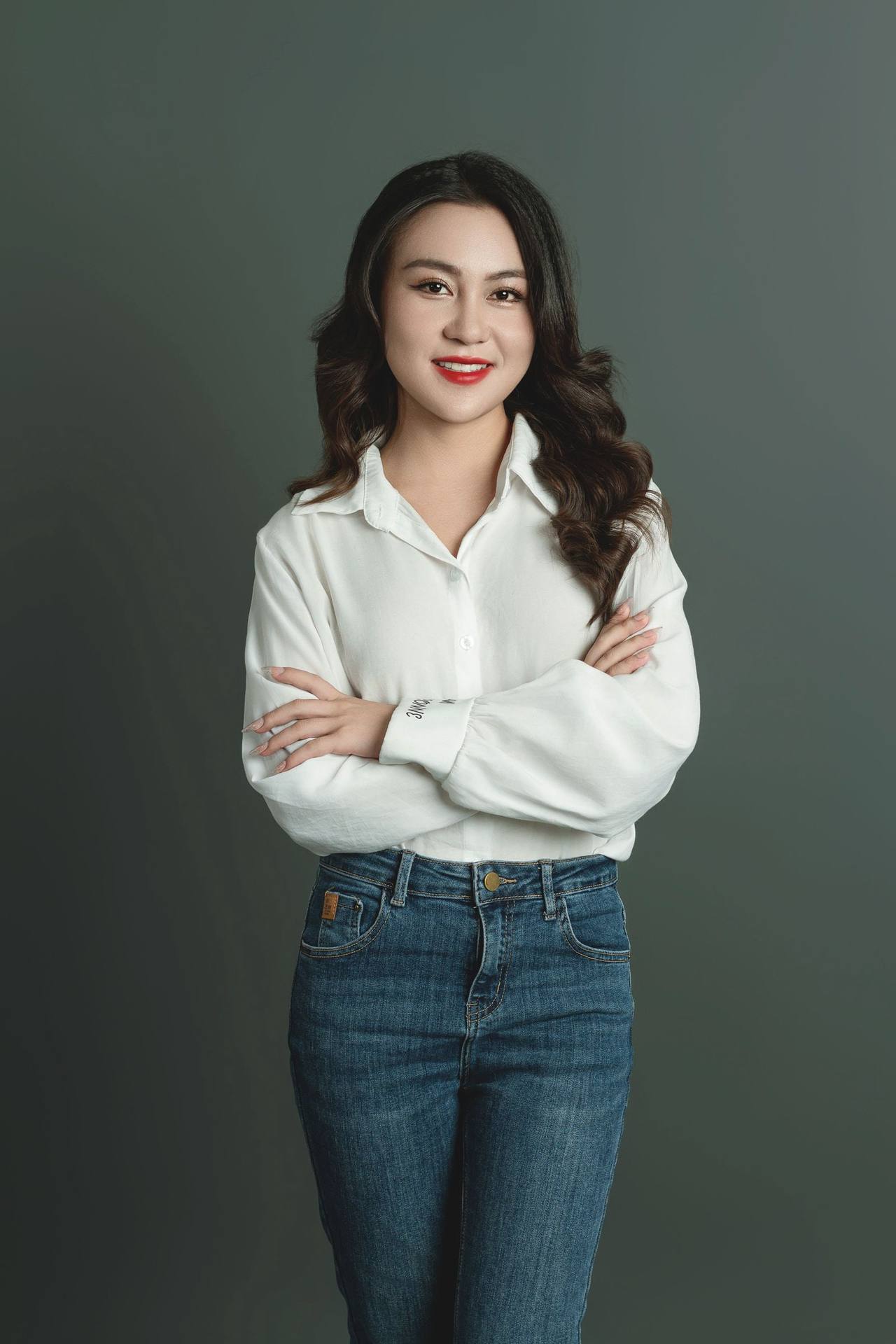 Chân dung người phụ nữ Việt Nam mặc áo sơ mi trắng và quần jeans, khoanh tay mỉm cười trước phông nền xám xanh