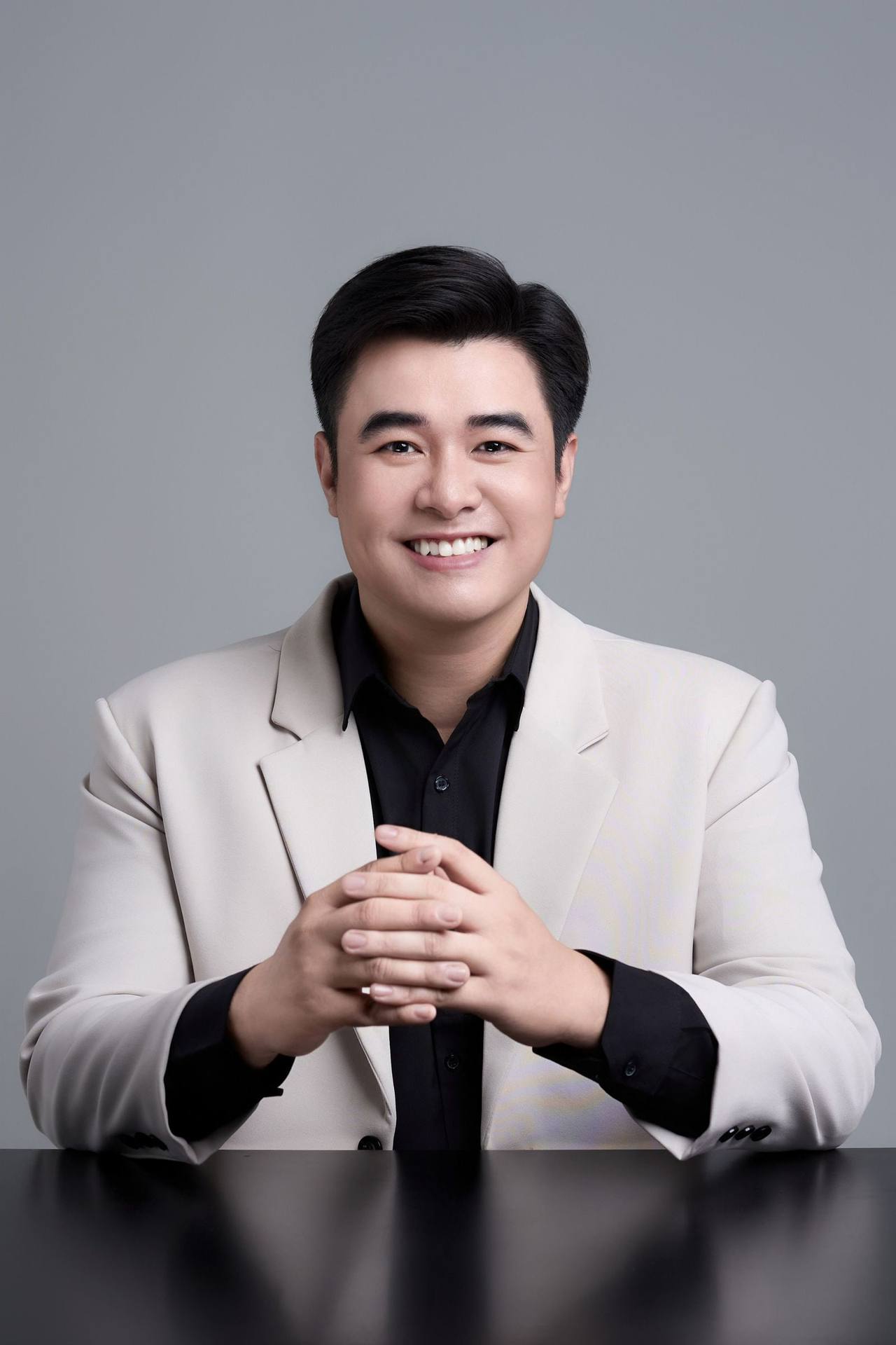 Chân dung người đàn ông Việt Nam mặc áo vest màu kem, ngồi đan tay trên bàn và mỉm cười trước phông nền xám
